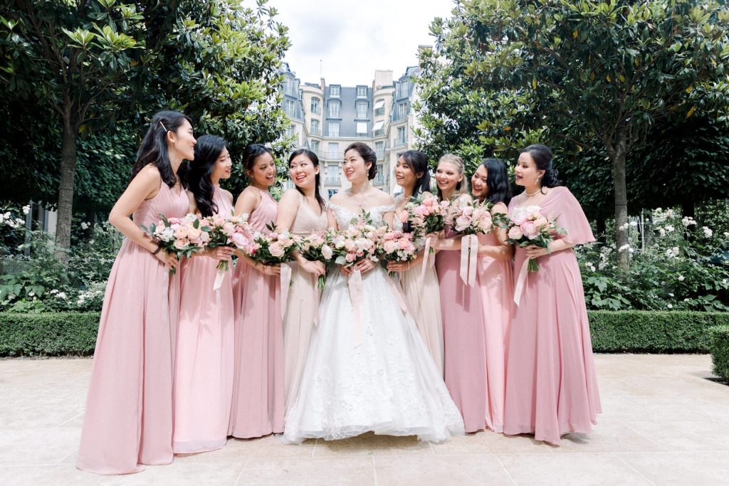 Mariée posée avec ses demoiselles d'honneur en robes roses tenant des bouquets dans un jardin parisien
