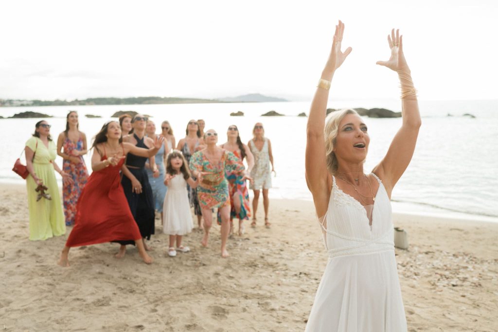 Mariée levant les bras pour lancer son bouquet aux invitées rassemblées sur une plage en Bretagne