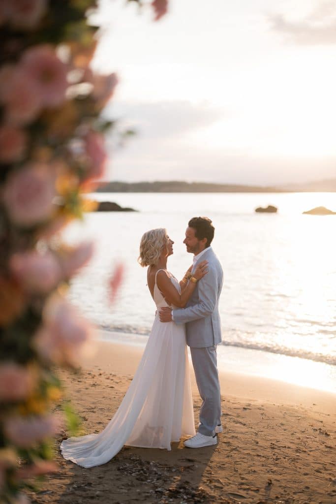 Mariés s'enlaçant face à la mer au coucher de soleil sous une arche florale sur la plage