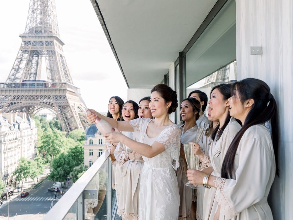 The bride opens a bottle of champagne on a balcony with her bridesmaids as the iconic Eiffel Tower rises in the background.