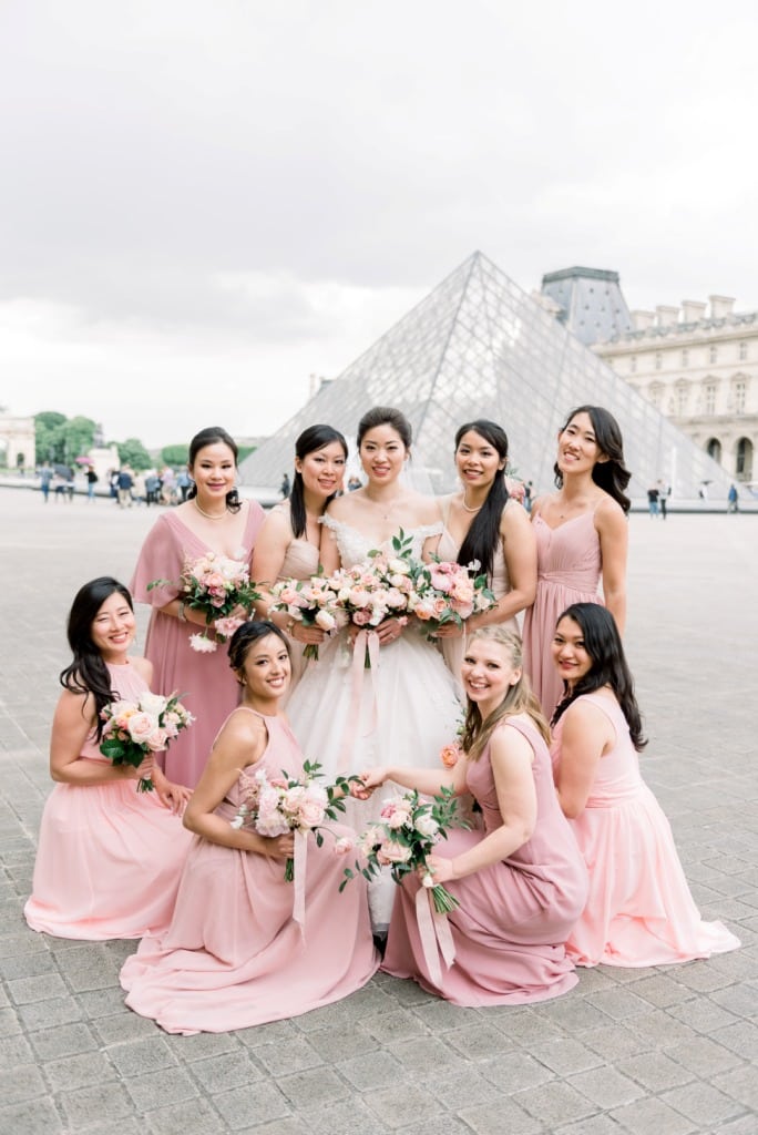 The bride and her bridesmaids pose together for a group photo in front of the iconic Louvre Museum in Paris.